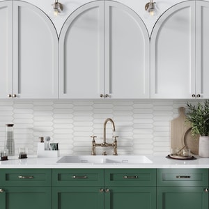 May include: A white kitchen with arched cabinets and green lower cabinets. The countertop is white with a double sink and a brass faucet. The backsplash is white subway tile.