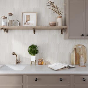May include: A white kitchen with a white subway tile backsplash, a wooden shelf with a plant, a picture frame, and a glass jar. The kitchen also has a stainless steel sink and a brown countertop.