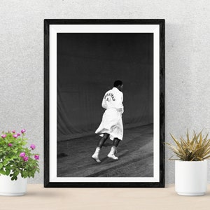 May include: Black and white photograph of a man wearing a white robe with "Muhammad Ali" printed on the back. The man is walking away from the camera.