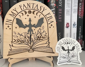 May include: A wooden bookend with a black dragon and floral design. The bookend is shaped like an arch and has the text "In My Fantasy Era" on it. The bookend is on a stand and there is a sticker with the same design on the stand.