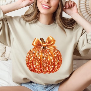May include: A beige sweatshirt featuring a sparkling orange pumpkin design with a bow. The pumpkin is covered in glitter and has a brown stem. The sweatshirt is a fall-themed item, perfect for the autumn season.