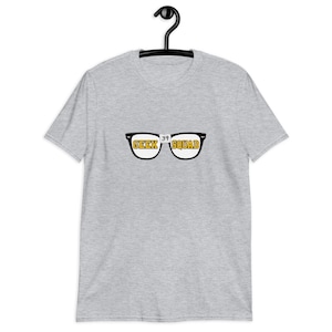 May include: A light grey t-shirt hanging on a black hanger. The shirt displays a graphic of spectacles with the words "GEEK SQUAD" in yellow. The number "39" is positioned between the lenses.