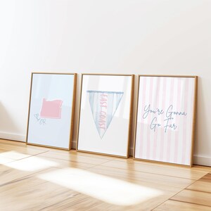 Op de afbeelding: Drie ingelijste kunstdrukken met gouden frames. De prints tonen een roze omtrek van de staat Oregon, een blauwe wimpel met de tekst "East Coast" en roze en witte strepen met de tekst "You're Gonna Go Far".
