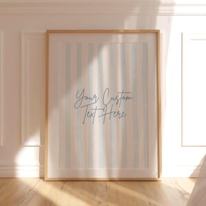 Op de afbeelding: Een ingelijste print met een licht houten frame. Het kunstwerk heeft verticale strepen in blauwe, beige en witte tinten. De tekst "Your Custom Text Here" is in cursief geschreven. Het frame leunt tegen een witte muur.