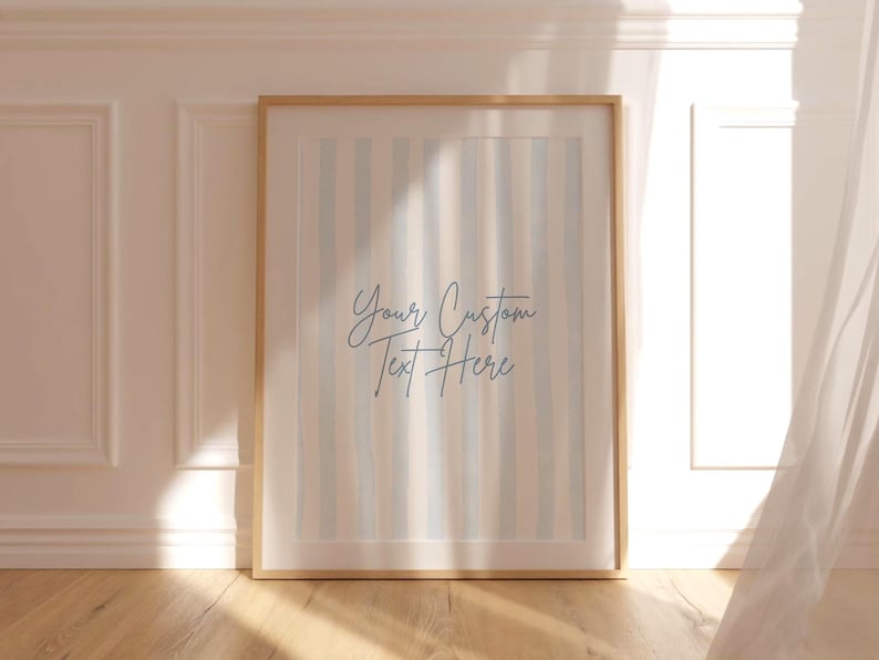 Op de afbeelding: Een ingelijste print met een licht houten frame. Het kunstwerk heeft verticale strepen in blauwe, beige en witte tinten. De tekst "Your Custom Text Here" is in cursief geschreven. Het frame leunt tegen een witte muur.
