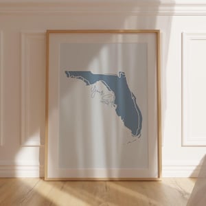 Op de afbeelding: Een ingelijste print met een blauwe omtrek van de staat Florida op een witte achtergrond. Het woord "Yours" is in cursief geschreven. De lijst is licht van kleur en de print staat tegen een witte muur.