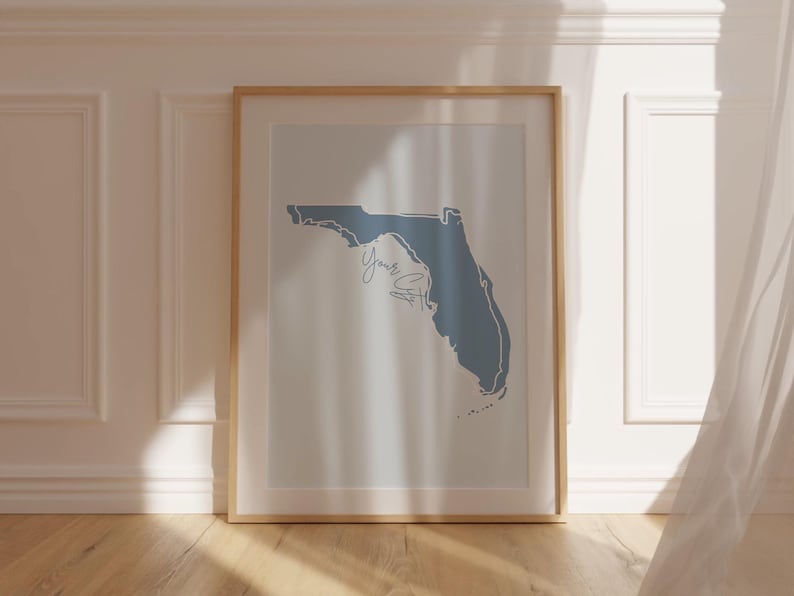 Op de afbeelding: Een ingelijste print met een blauwe omtrek van de staat Florida op een witte achtergrond. Het woord "Yours" is in cursief geschreven. De lijst is licht van kleur en de print staat tegen een witte muur.