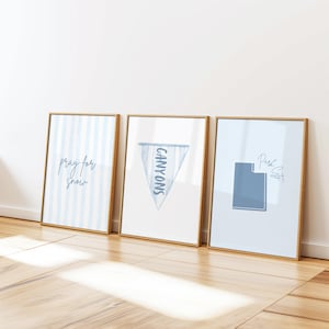Op de afbeelding: Drie ingelijste kunstdrukken met goudkleurige frames. De prints tonen blauw-witte ontwerpen, waaronder verticale strepen, een wimpel met de tekst "CANYONS" en een Utah-omtrek met de tekst "Park City". De prints leunen tegen een witte muur.
