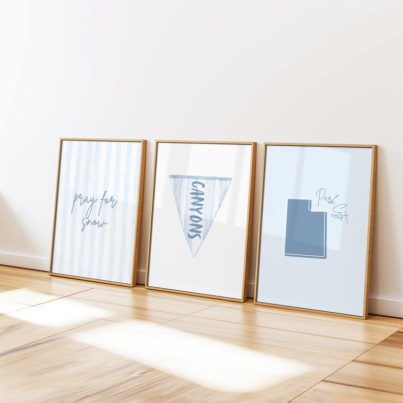 Op de afbeelding: Drie ingelijste kunstdrukken met goudkleurige frames. De prints tonen blauw-witte ontwerpen, waaronder verticale strepen, een wimpel met de tekst "CANYONS" en een Utah-omtrek met de tekst "Park City". De prints leunen tegen een witte muur.