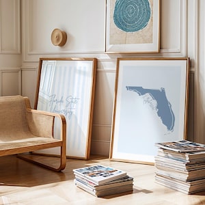 Op de afbeelding: Een lichte kamer met ingelijste kunst en een rieten stoel. Twee ingelijste prenten leunen tegen een muur, een met de tekst "Florida State University" en de andere een blauwe kaart van Florida. Een stapel tijdschriften ligt op de vloer.