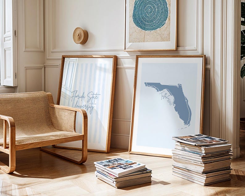 Op de afbeelding: Een lichte kamer met ingelijste kunst en een rieten stoel. Twee ingelijste prenten leunen tegen een muur, een met de tekst "Florida State University" en de andere een blauwe kaart van Florida. Een stapel tijdschriften ligt op de vloer.