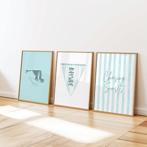 Op de afbeelding: Drie ingelijste kunstdrukken met gouden frames leunen tegen een witte muur. De prints tonen een kaart van Maryland, een wimpel met "Bayside" en blauw-witte strepen met de tekst "Chasing Sunsets". De prints staan op een houten vloer.