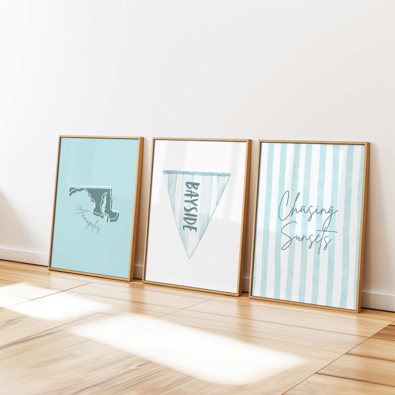 Op de afbeelding: Drie ingelijste kunstdrukken met gouden frames leunen tegen een witte muur. De prints tonen een kaart van Maryland, een wimpel met "Bayside" en blauw-witte strepen met de tekst "Chasing Sunsets". De prints staan op een houten vloer.
