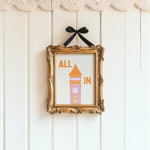 May include: A gold framed print with the text "ALL IN" and a cartoon illustration of a purple clock tower with an orange roof.