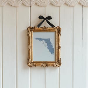 Op de afbeelding: Een ingelijste print van de blauwe omtrek van Florida tegen een lichtblauwe achtergrond. De sierlijke gouden lijst wordt opgehangen door een zwarte strik aan een witte wand met panelen. Het woord "Tallahassee" is zichtbaar.