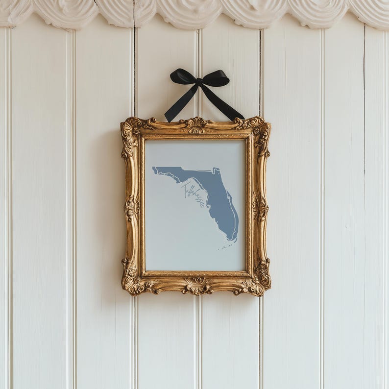 Op de afbeelding: Een ingelijste print van de blauwe omtrek van Florida tegen een lichtblauwe achtergrond. De sierlijke gouden lijst wordt opgehangen door een zwarte strik aan een witte wand met panelen. Het woord "Tallahassee" is zichtbaar.