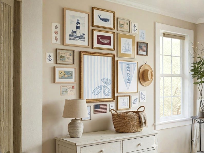 Op de afbeelding: Een galerijmuur met ingelijste nautische kunst, waaronder een vuurtoren, walvissen en zeilboten. Een geweven mand, een lamp en een strohoed maken de kustdecoratie compleet. Het kleurenpalet omvat blauw, wit en beige.