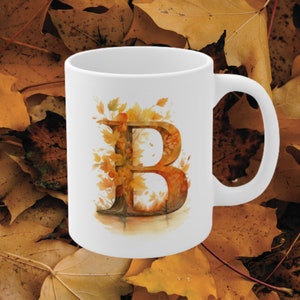 May include: White ceramic mug with a watercolor illustration of the letter B surrounded by autumn leaves in shades of orange, yellow, and brown.