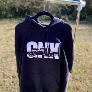 Op de afbeelding: Zwart hoodie met een witte afbeelding van een auto en het woord "G.O.A.T." gedrukt op de voorkant.