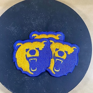 Könnte beinhalten: Zwei gestickte Bären-Patches in Blau und Gold. Die Bären sind nach vorne gerichtet, mit offenem Maul und sichtbaren Zähnen. Die Patches befinden sich auf einer schwarzen Oberfläche.