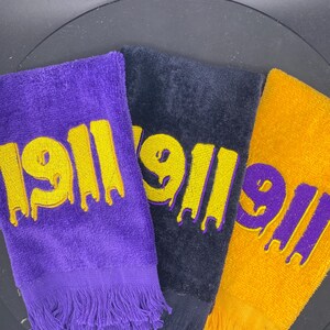 Puede incluir: Tres toallas de rizo en morado, negro y dorado. Cada toalla tiene el número "1911" bordado en un color contrastante con un efecto de goteo. Las toallas tienen bordes con flecos.