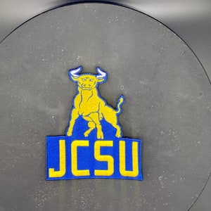 May include: An embroidered patch featuring a yellow bull mascot with the text "JCSU" in yellow lettering on a blue background.