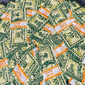 Könnte beinhalten: Nahaufnahme einer Stapel von grünen und weißen gestickten Patches in Form von Geldstapeln. Die Patches haben orange und weiße Nähte an den Rändern.