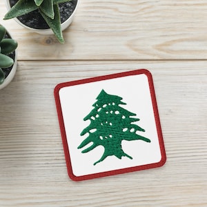 May include: A white square coaster with a red border features a green cedar tree, the national symbol of Lebanon.