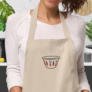 Puede incluir: Un delantal beige con tirantes ajustables y un bolsillo con un diseño bordado de un cuenco colorido con flores y hojas estilizadas.