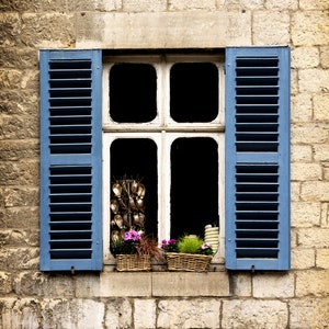 May include: A blue shuttered window with white trim and a view of a flower pot with pink flowers. The window is set in a stone wall.