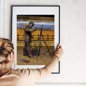 May include: A framed photograph of a bronze statue of a person painting with a brush in front of a building with a blurred light trail in the background.