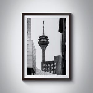 Könnte beinhalten: Ein Schwarzweißfoto eines hohen Turms mit einer spitzen Spitze, umgeben von Gebäuden. Der Turm befindet sich in der Mitte des Bildes und ist das höchste Bauwerk. Die Gebäude befinden sich auf beiden Seiten des Turms und sind niedriger als der Turm. Das Foto wurde aus einer niedrigen Perspektive aufgenommen und zeigt den Turm von unten.