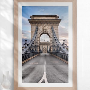 May include: A black and white photograph of the Chain Bridge in Budapest, Hungary. The bridge is a suspension bridge with a stone archway at the center. The road leading to the bridge is empty and the sky is a light blue.