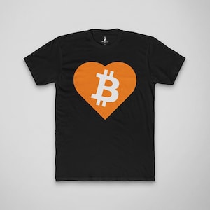 May include: Black t-shirt featuring a large orange heart with a white Bitcoin symbol in the center. The Bitcoin symbol is a stylized letter "B" with two vertical lines. The shirt is a classic crew neck style.