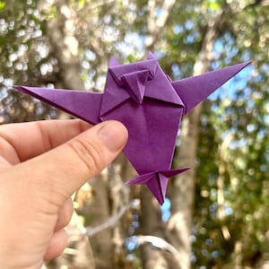Puede incluir: Un pájaro de origami púrpura con las alas extendidas se sostiene en una mano contra un fondo borroso de follaje verde. El pájaro de papel tiene un pico y una cola puntiagudos, y las alas están muy anguladas.