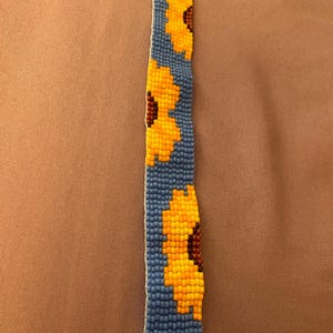 May include: A beaded bracelet featuring a blue background with three yellow and brown sunflower designs. The bracelet has gold-colored metal ends. The beadwork is detailed and the sunflowers are in full bloom.