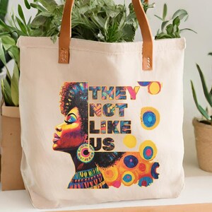 Op de afbeelding: Een beige canvas draagtas met bruine leren handvatten. De tas heeft een kleurrijke afbeelding van het profiel van een vrouw met de tekst "THEY NOT LIKE US". Het portret van de vrouw is in tinten paars, geel en blauw.