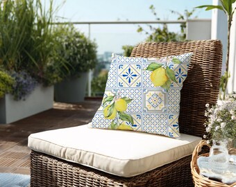 Könnte beinhalten: Quadratisches Zierkissen mit Zitronen- und Fliesenmuster. Das Kissen zeigt ein Aquarelldesign von Zitronen und grünen Blättern auf einem blau-weißen Fliesenmuster. Das Kissen liegt auf einem Korbstuhl mit cremefarbenem Kissen.