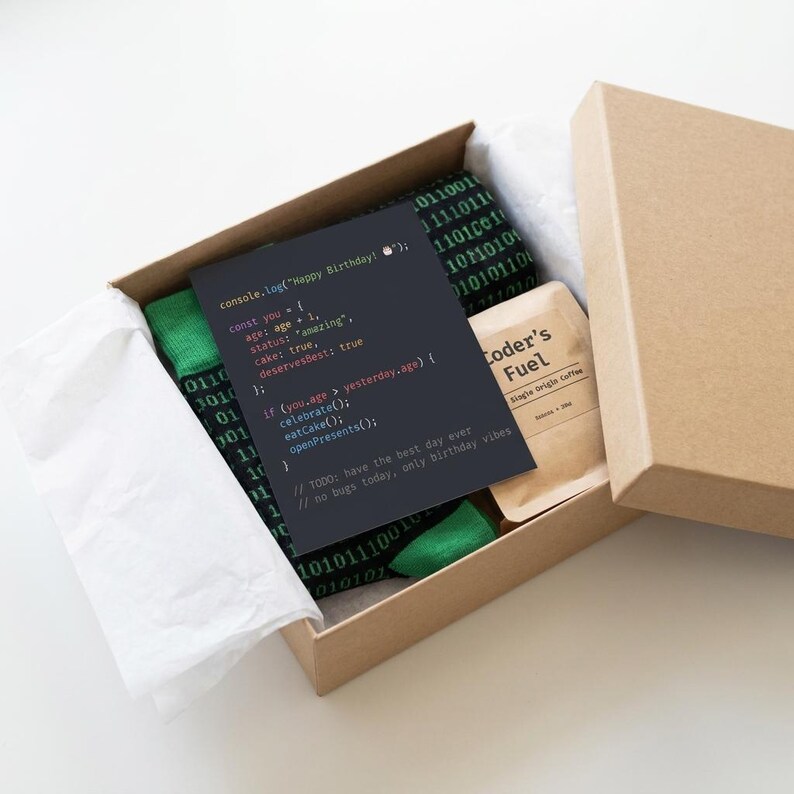 K&ouml;nnte beinhalten: Eine Geschenkschachtel aus Karton mit gr&uuml;nen Socken mit schwarzem Bin&auml;rcode-Muster, einem kleinen braunen Beutel mit der Aufschrift Coder's Fuel und einer dunkelgrauen Karte mit wei&szlig;em und farbigem Text, der console.log(Happy Birthday); lautet.