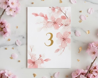 May include: A rectangular table number card featuring the number 3 in a gold serif font, centered on a white background. The card is decorated with watercolor-style illustrations of light pink cherry blossom branches and scattered gold flakes.