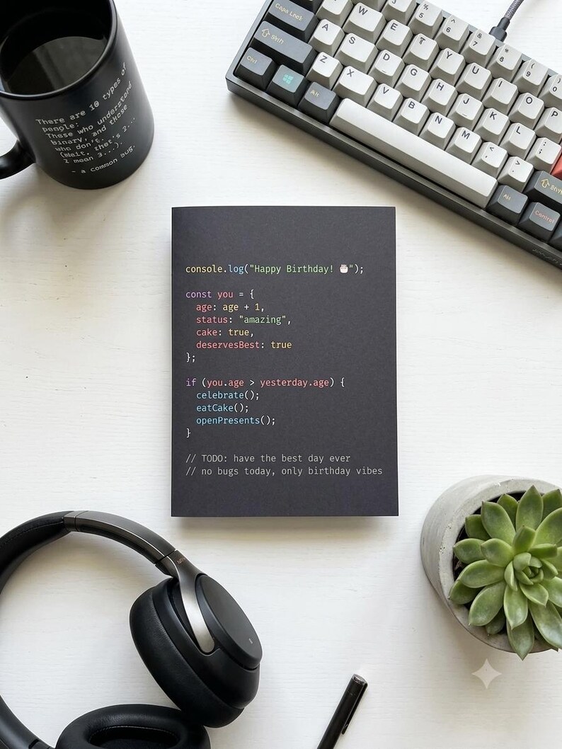 K&ouml;nnte beinhalten: Eine schwarze rechteckige Gru&szlig;karte mit farbigem Programmiercode-Text: console.log("Happy Birthday! 🎂"); const you = { age: age + 1, status: "amazing", cake: true, deservesBest: true }; if (you.age > yesterday.age) { celebrate(); eatCake(); openPresents(); } // TODO: have the best day ever // no bugs today, only birthday vibes