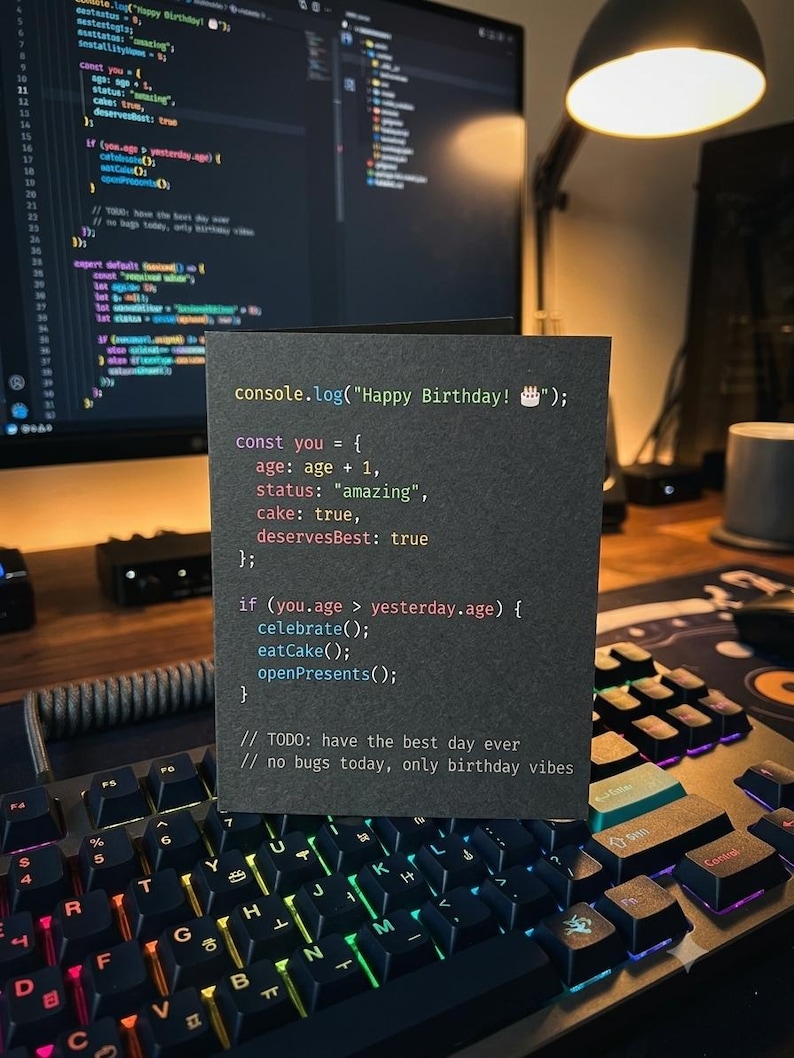K&ouml;nnte beinhalten: Eine schwarze Gru&szlig;karte mit farbigem Programmiercode-Text, der lautet: console.log("Happy Birthday! 🎂"); const you = { age: age + 1, status: "amazing", cake: true, deservesBest: true }; if (you.age > yesterday.age) { celebrate(); eatCake(); openPresents(); } // TODO: have the best day ever // no bugs today, only birthday vibes