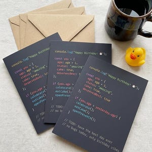 K&ouml;nnte beinhalten: Drei dunkelgraue Gru&szlig;karten mit wei&szlig;em, rotem und blauem Computercode-Text, der console.log("Happy Birthday!") lautet, zusammen mit einer gelben Gummiente und einer dunkelbraunen Keramiktasse. Die Karten werden mit hellbraunen Umschl&auml;gen geliefert.
