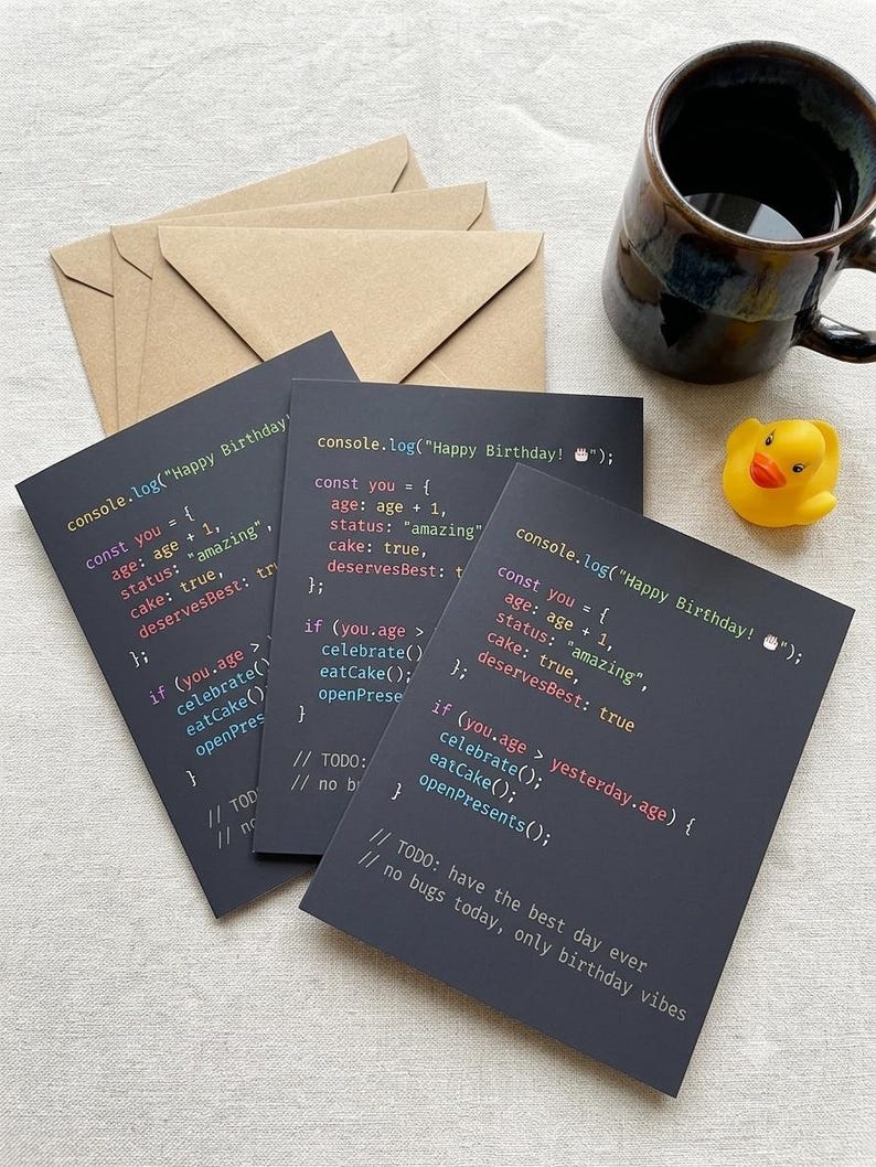 K&ouml;nnte beinhalten: Drei dunkelgraue Gru&szlig;karten mit wei&szlig;em, rotem und blauem Computercode-Text, der console.log("Happy Birthday!") lautet, zusammen mit einer gelben Gummiente und einer dunkelbraunen Keramiktasse. Die Karten werden mit hellbraunen Umschl&auml;gen geliefert.