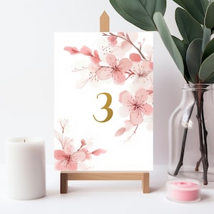 May include: A white card with a pink floral design and the number 3 in gold. The card is on a small wooden easel.