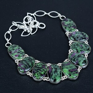 May include: A silver necklace featuring oval-shaped green and purple stones. The stones are set in a silver bezel and connected by a silver chain. The necklace is displayed on a black surface.