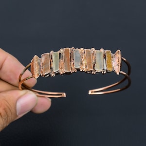 Może przedstawiać: Miedziana bransoletka na nadgarstek z rzędem prostokątnych, wielokolorowych kamieni. Kamienie są w odcieniach beżu, brązu i szarości. Bransoletka ma prosty, otwarty design.