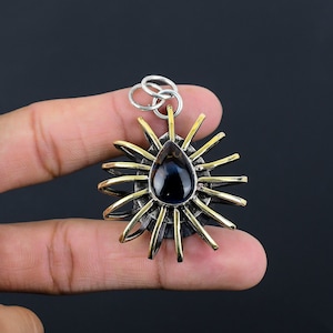 May include: A silver and gold pendant with a dark blue, teardrop-shaped stone at its center. The pendant has a sunburst design with gold rays extending from the central stone. The pendant is held by a person's fingers against a dark background.