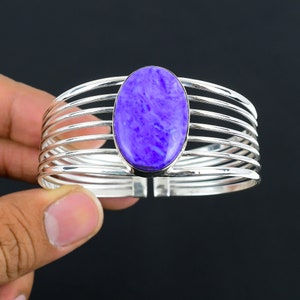 Pode incluir: Uma pulseira de punho prateada com uma grande pedra preciosa oval roxa. A pulseira tem um design de várias bandas com uma moldura central para a pedra. A pedra preciosa tem uma aparência marmorizada.