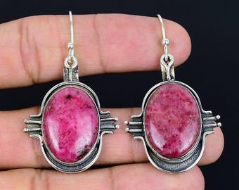 May include: A pair of silver earrings with oval, vibrant pink gemstones. The gemstones are set in a decorative silver frame. The earrings have a hook closure. The design is unique.